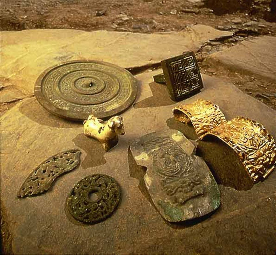 Erste Funde: ein goldener Armreif, daneben die Schablone; zwei bronzene Zierscheiben; eine glasierte Tierskulptur; ein Kultspiegel mit chinesischen Schriftzeichen; ein quadratisches Beamtensiegel