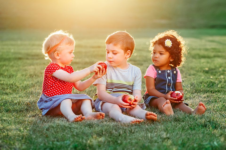 Drei Kinder sitzen auf einer Wiese und teilen Äpfel Drei Kinder sitzen auf einer Wiese und teilen Äpfel