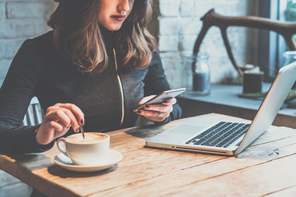 Frau sitzt beim Arbeiten am Laptop an einem Holztisch, trinkt Kaffee und benutzt ein Smartphone