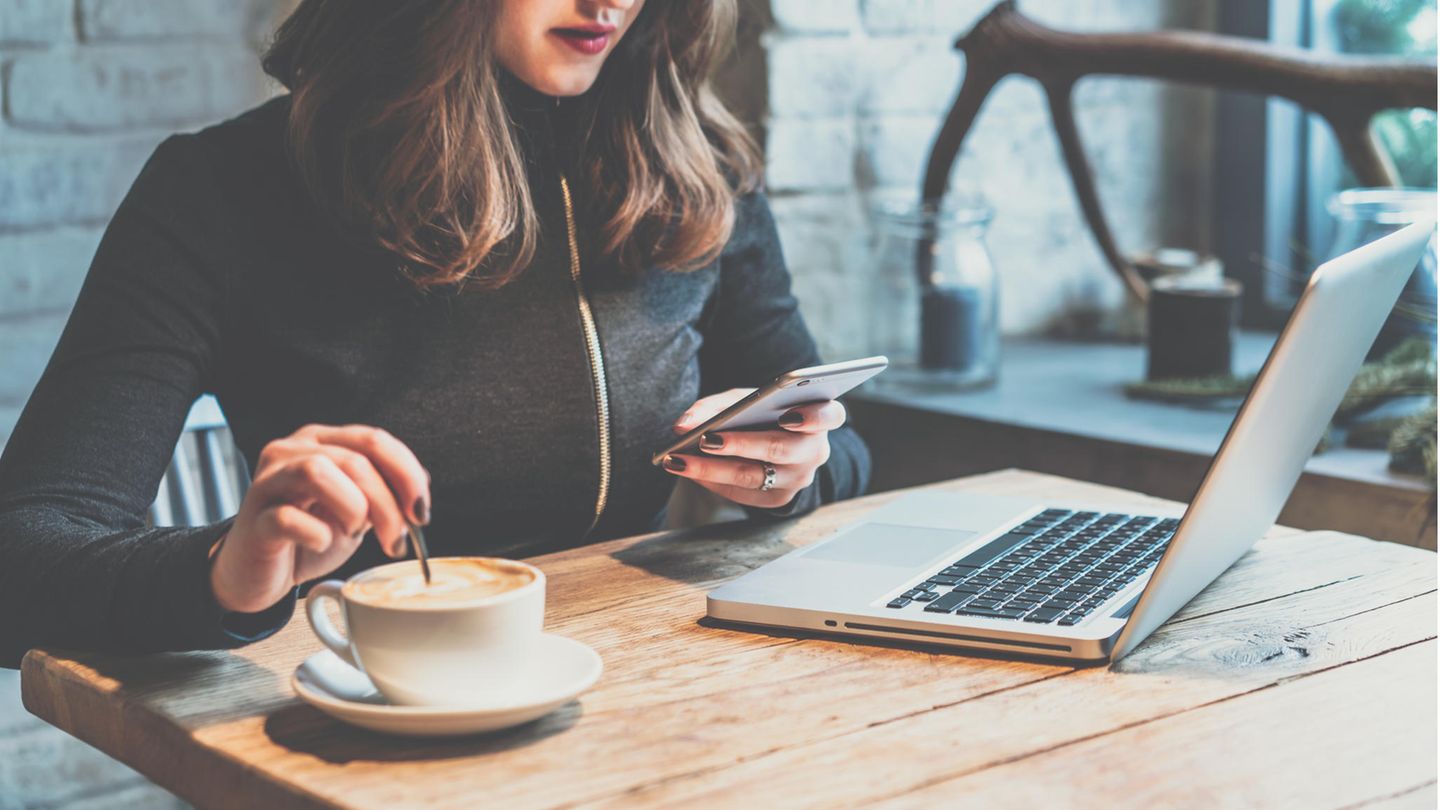 Frau sitzt beim Arbeiten am Laptop an einem Holztisch, trinkt Kaffee und benutzt ein Smartphone