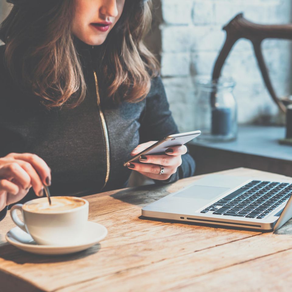 Frau sitzt beim Arbeiten am Laptop an einem Holztisch, trinkt Kaffee und benutzt ein Smartphone