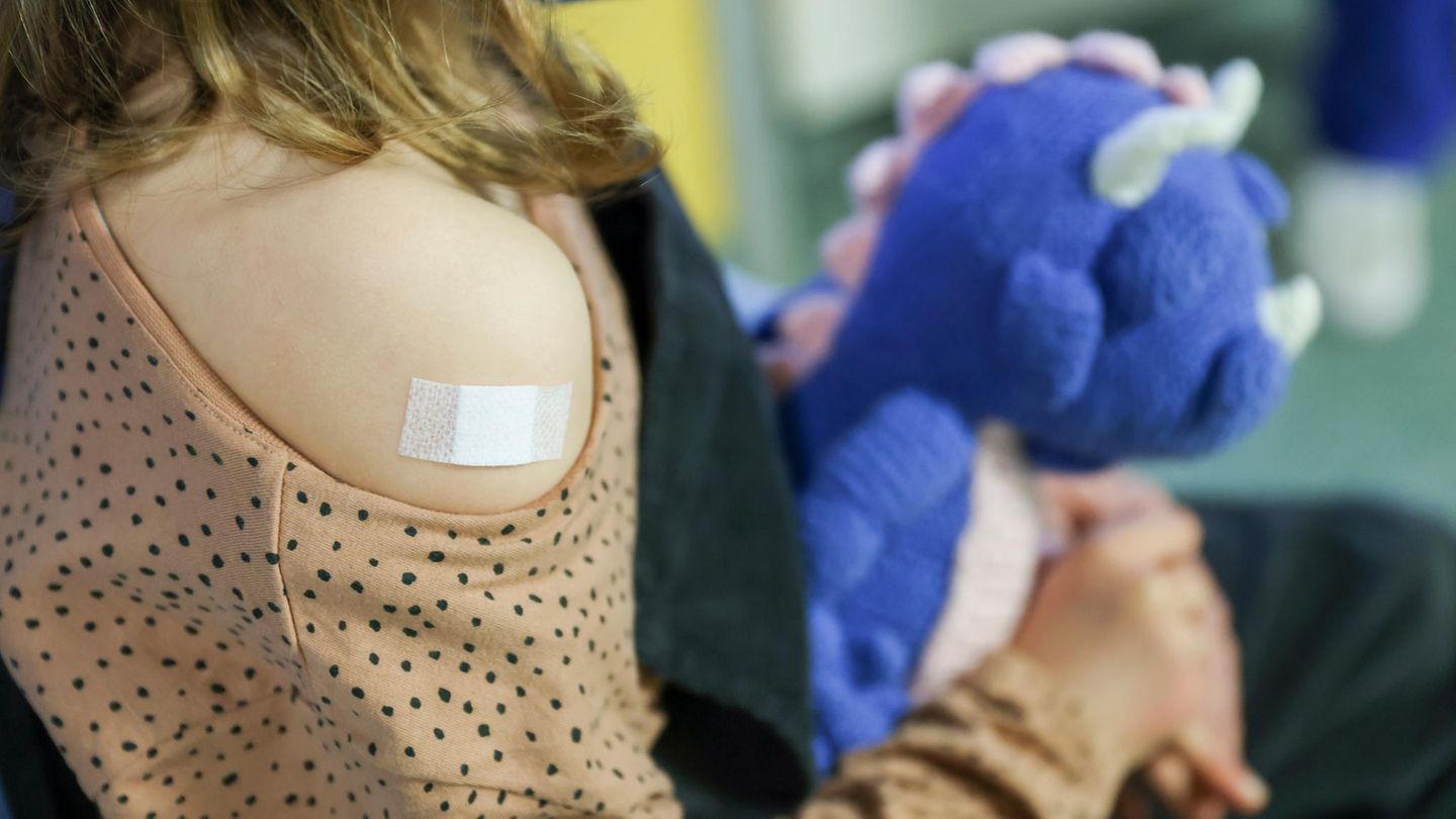 KInd sitzt mit einem Pflaster auf dem Arm und Kuscheltier in der Hand in einer Arztpraxis