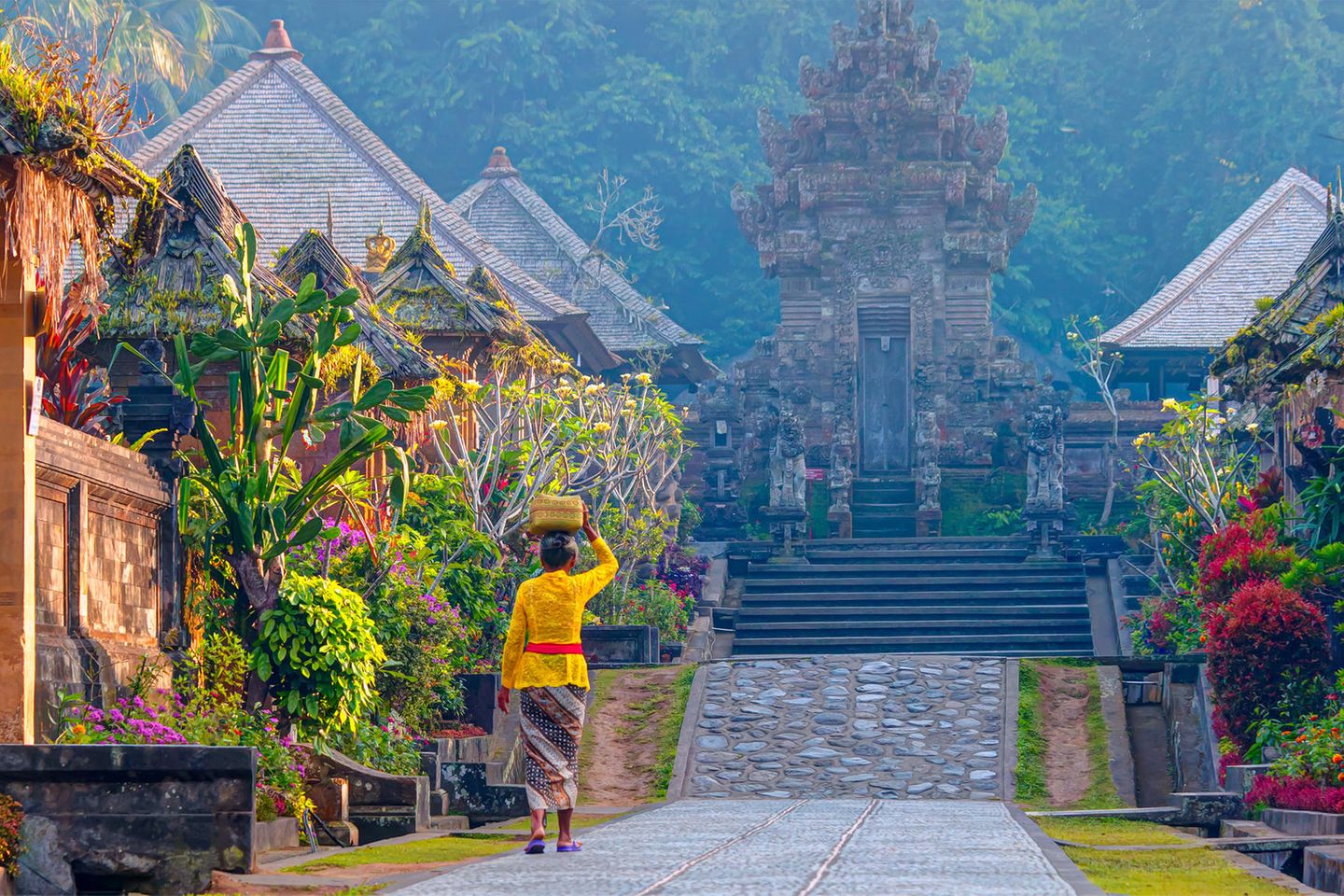 Eine bunt gekleidete Frau läuft auf einen Tempel in einem balinesischen Dorf zu