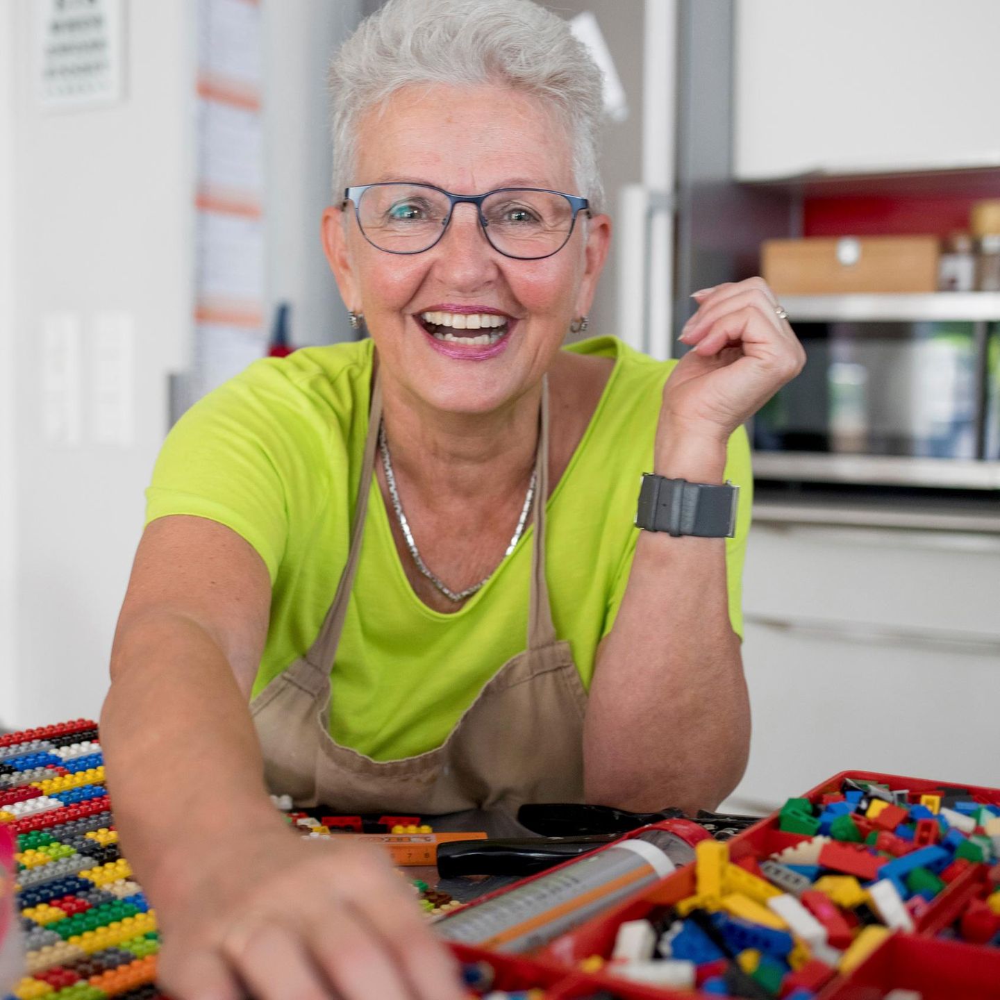 Eine ältere Frau sitzt in ihrer Küche und hat viele Legosteine vor sich auf dem Tisch liegen