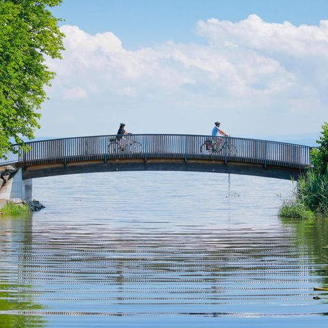 Zwei Fahrradfahrer überqueren die Brücke über den Bach Aach bei der Mündung in den Bodensee