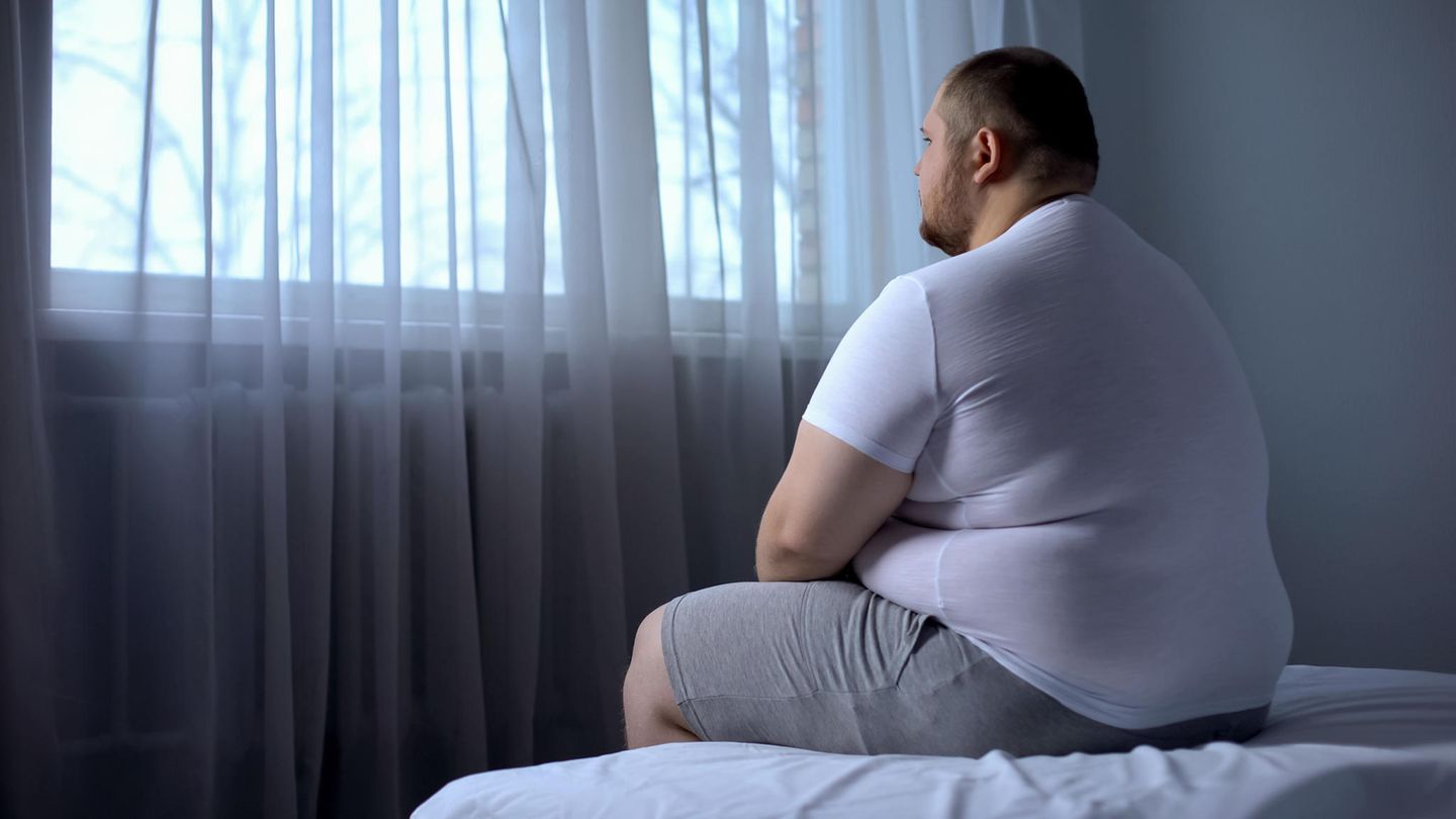 Ein übergewichtiger Mann sitzt auf einem Bett und schaut aus dem Fenster