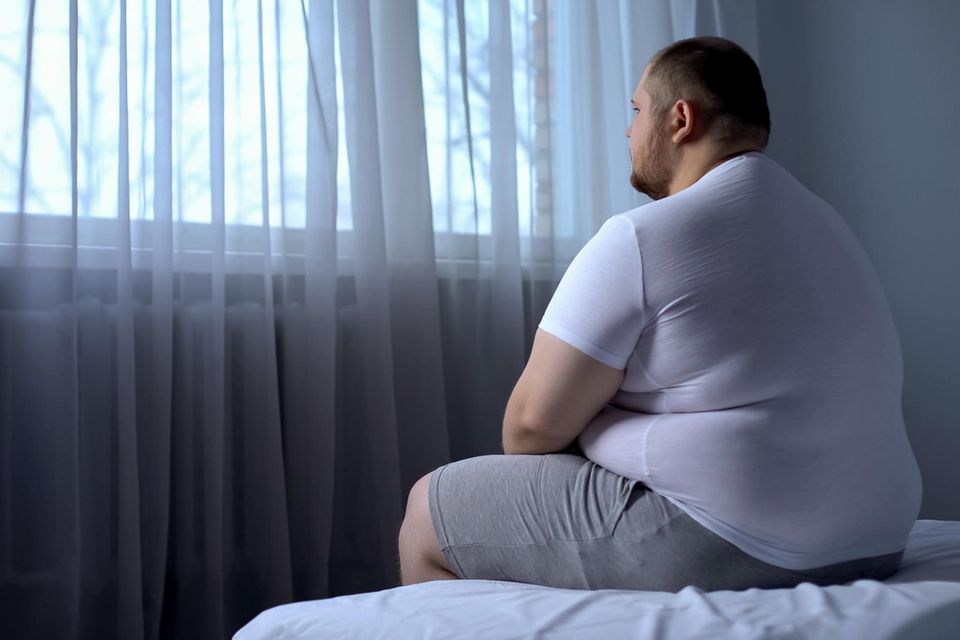 Ein übergewichtiger Mann sitzt auf einem Bett und schaut aus dem Fenster