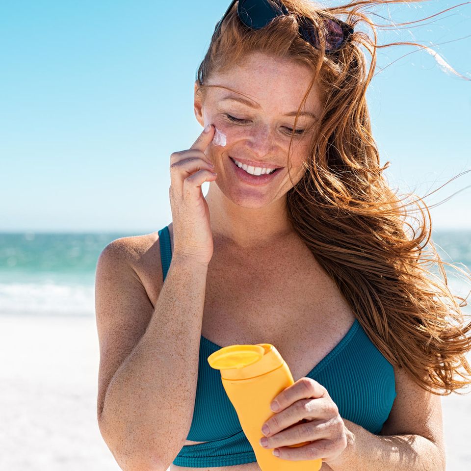 Eine rothaarige Frau cremt sich am Strand mit Sonnencreme ein