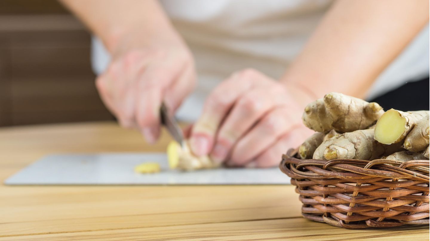 Weidenkörbchen mit Ingwerwurzeln, im Hintergrund schneidet eine Frau ein Stück Ingwer in feine Scheiben