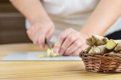 Weidenkörbchen mit Ingwerwurzeln, im Hintergrund schneidet eine Frau ein Stück Ingwer in feine Scheiben