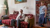 Ein älterer Mann sitzt auf einem Sofa, eine ältere Frau steht daneben und schaut den Mann an