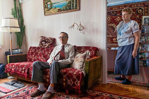 Ein älterer Mann sitzt auf einem Sofa, eine ältere Frau steht daneben und schaut den Mann an