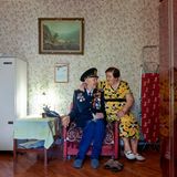 Ein alter Mann sitzt in einem Sessel, er trägt eine Uniform. Auf der Lehne des Sessels sitzt seine Frau