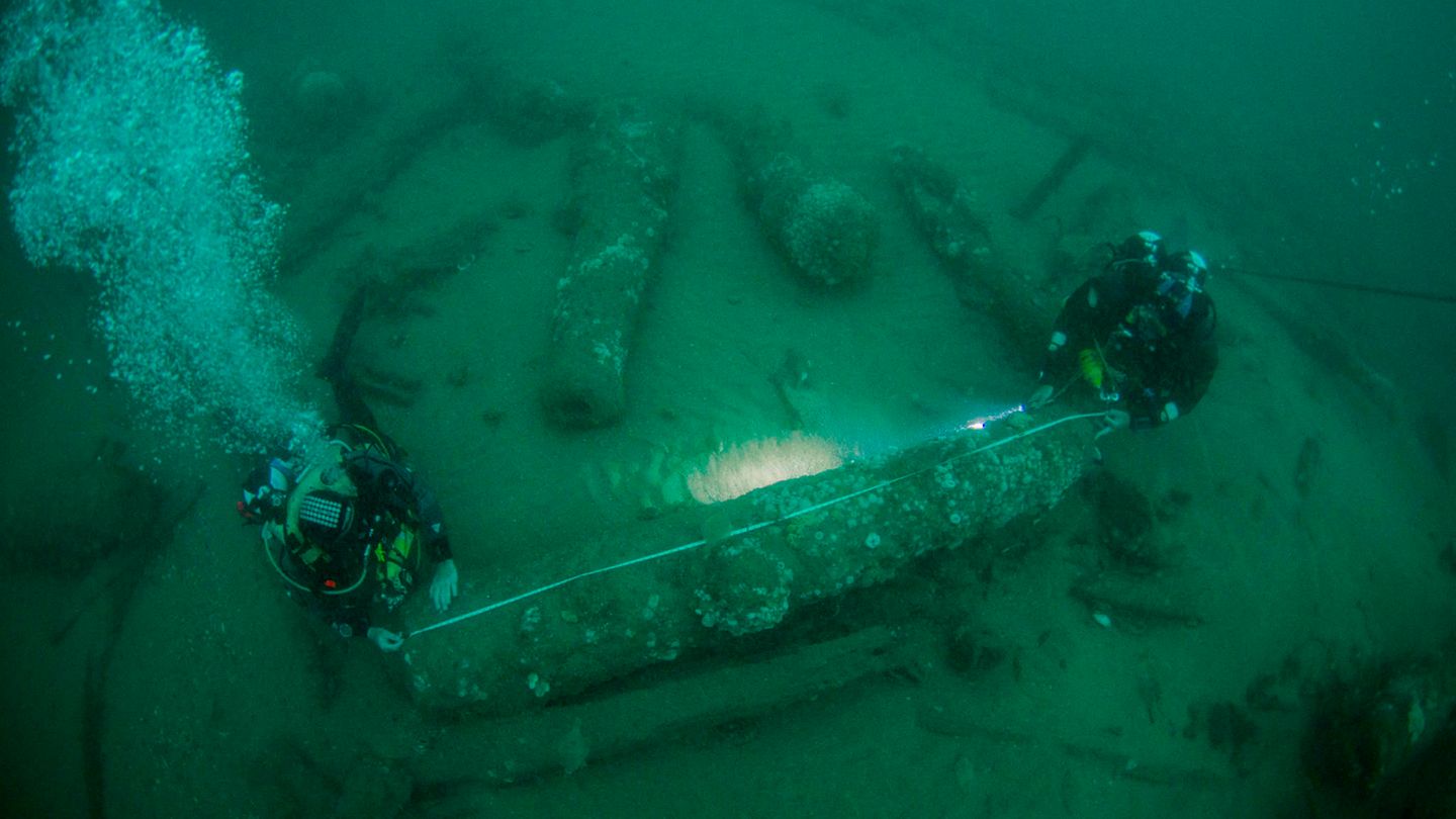 Zwei Taucher messen mit einem Maßband eine Kanone unter Wasser aus