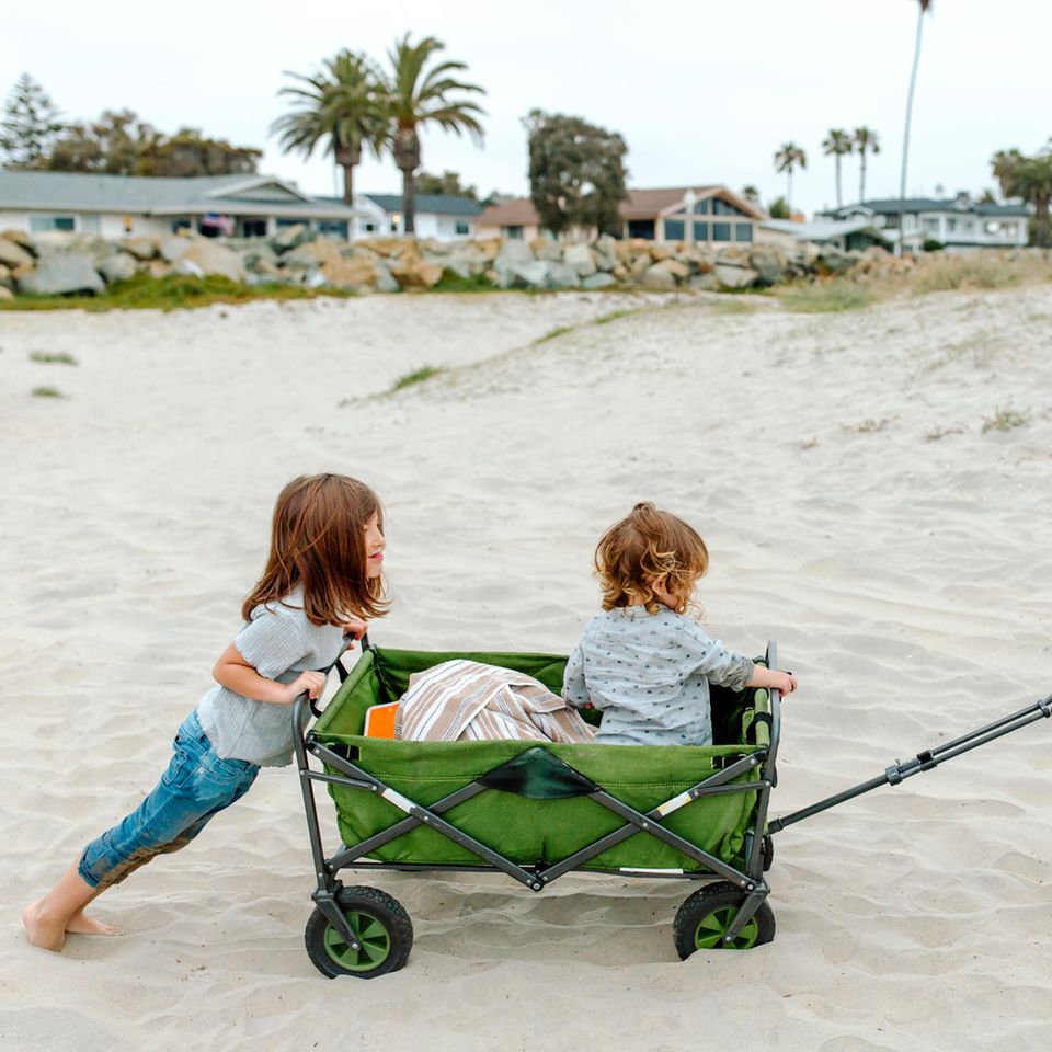 Drei Kinder mit einem faltbaren Bollerwagen am Strand.