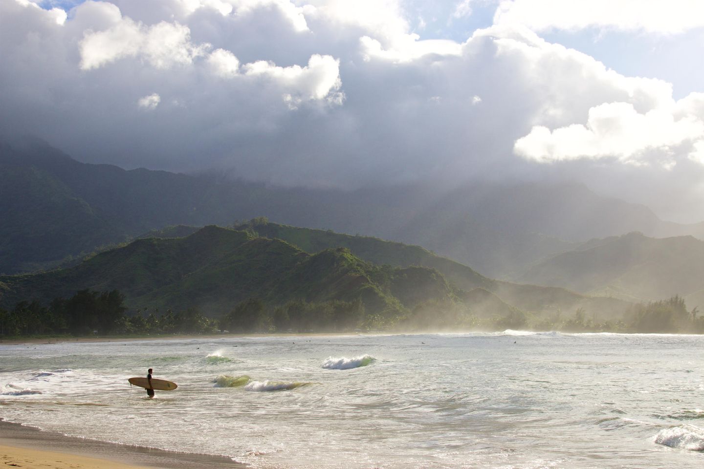 Surfen oder Paddeln? Ein Surfer geht mit seinem Brett ins Wasser. Im Hintergrund sind Berge