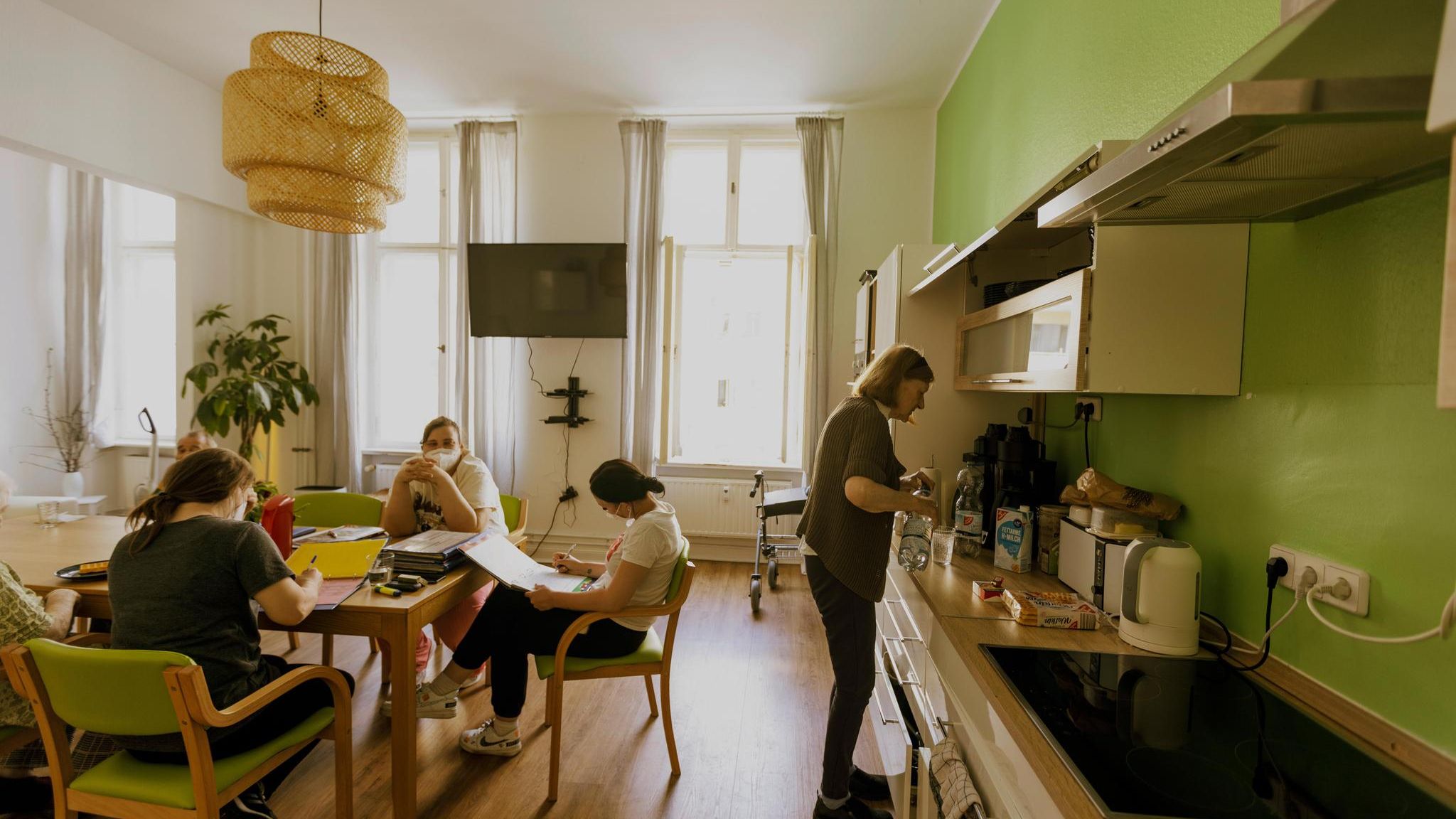 Die Küche in der Senior*innen WG in Berlin Charlottenburg. Am Tisch (links) gehen die Pflegerinnen die Mitbewohner*innen Akten durch. Rechts bereitet Frau Riechmann sich ein Getränk zu. Lebensmittelpunkt: Wie in jeder WG spielt sich der Großteil des Miteinanders in der Gemeinschaftsküche ab. Hier wird geplant, gekocht und gegessen. Es kommt, wer mag