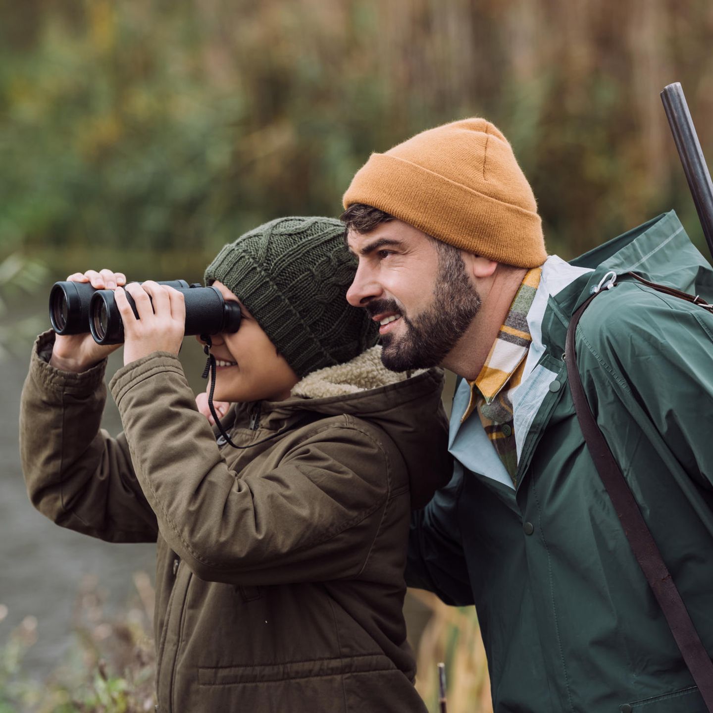 Ein Kind schaut durch ein Fernglas, daneben steht ein Mann mit einem Gewehr um die Schulter gehängt