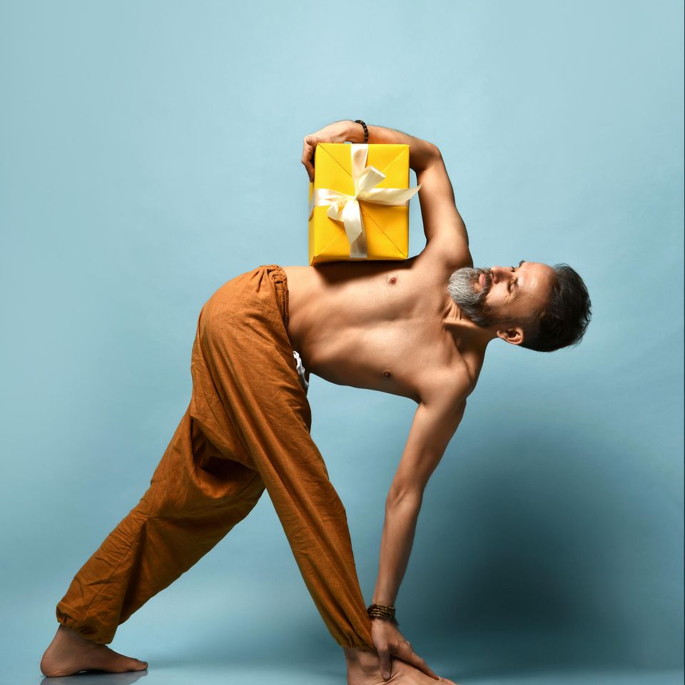 Geschenke für YogaYoga-Geschenke: Ein Mann nimmt eine Yogahaltung ein und hält dabei eine gelbe Geschenkschachtel unter dem Arm.