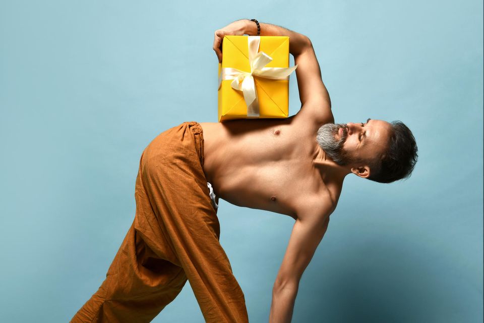 Geschenke für YogaYoga-Geschenke: Ein Mann nimmt eine Yogahaltung ein und hält dabei eine gelbe Geschenkschachtel unter dem Arm.