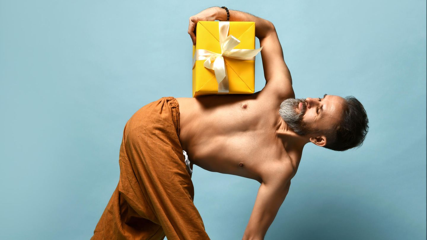 Geschenke für YogaYoga-Geschenke: Ein Mann nimmt eine Yogahaltung ein und hält dabei eine gelbe Geschenkschachtel unter dem Arm. Geschenke für YogaYoga-Geschenke: Ein Mann nimmt eine Yogahaltung ein und hält dabei eine gelbe Geschenkschachtel unter dem Arm.