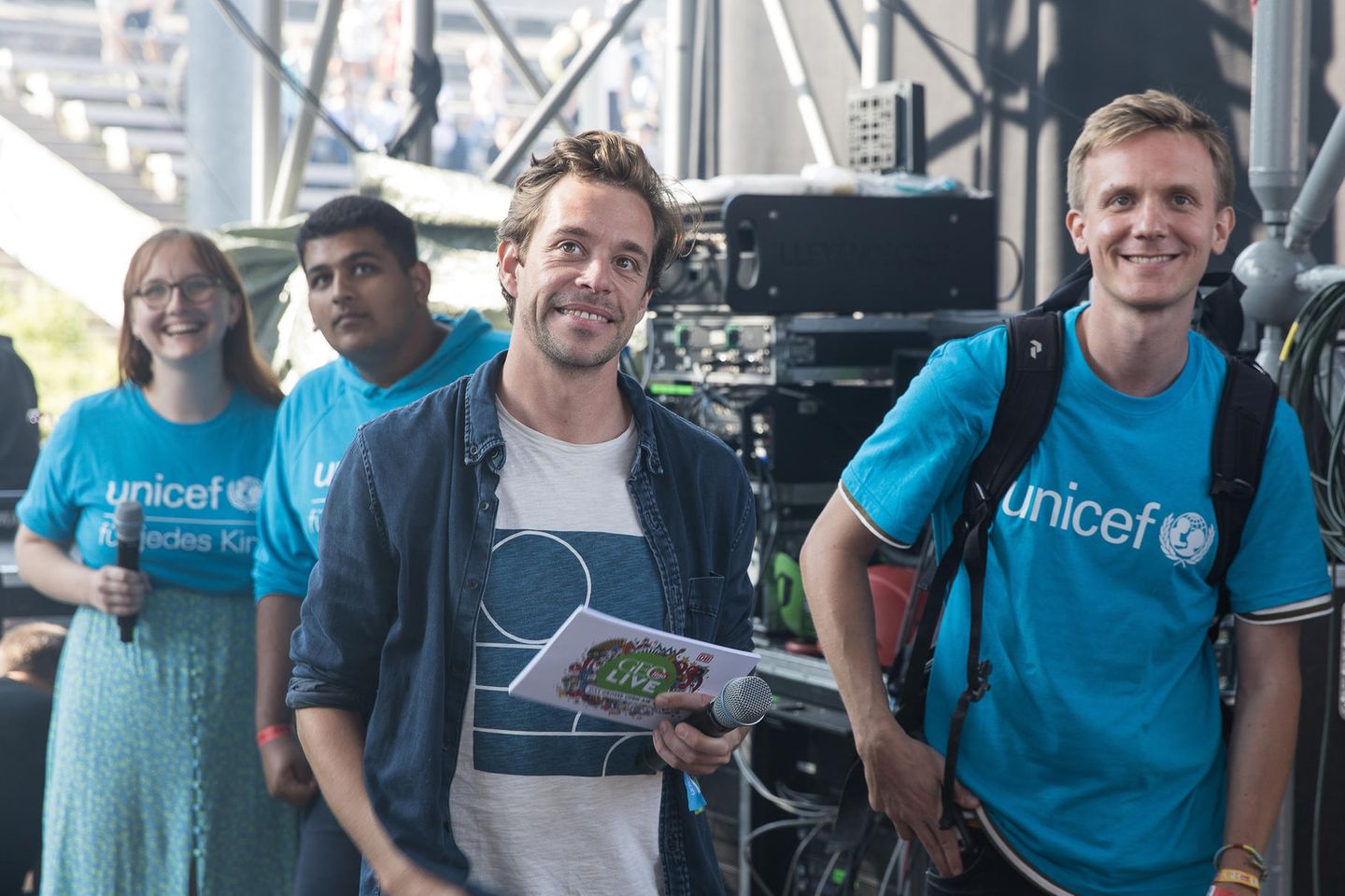 Tobias Krell alias "Cecker Tobi" (2. von rechts) steht mit Moderationskarten und einem Mikrofon hinter der Bühne.