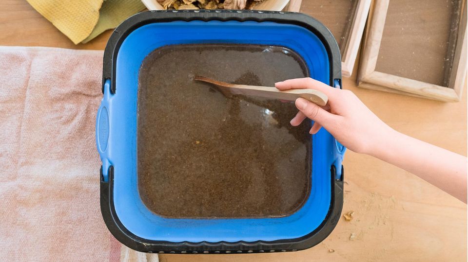 In einer quadratischen Wanne wird eine braune Flüssigkeit mit einem Kochlöffel umgerührt.