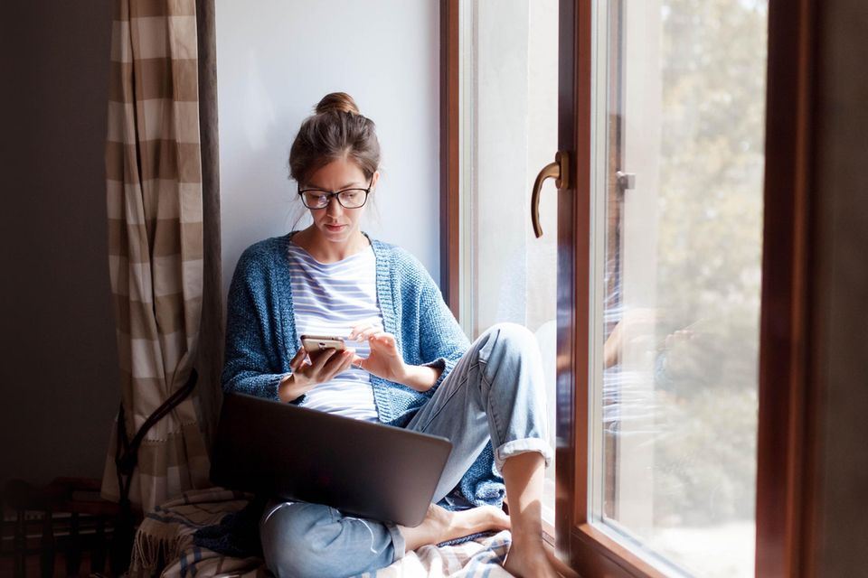 Eine junge Frau sitzt mit ihrem Laptop und Handy auf einer Fensterbank