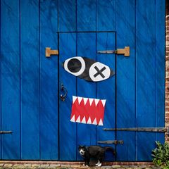 Eine blaue Tür, darauf sind Zähne und Augen aus Papier zu sehen. Eine schwarz-weiße Katze steht unten davor.
