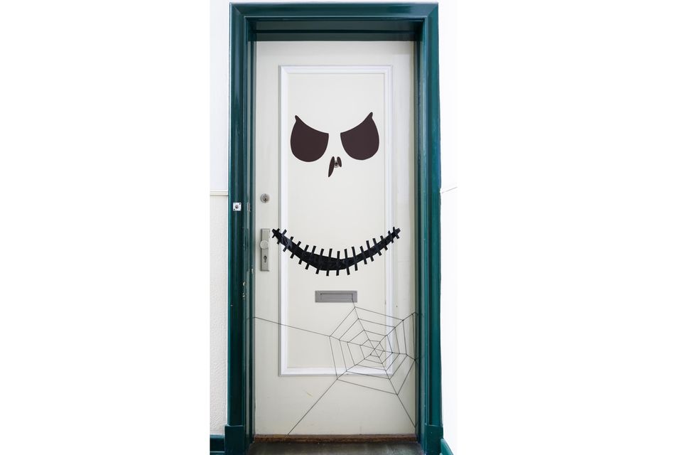 Eine weiße Tür. Darauf sind schwarze Geisteraugen, zwei Nasenlöcher und ein breiter Mund zu sehen, unten rechts ein Spinnennetz.