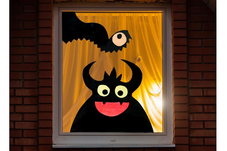 Ein Fenster von außen mit einem weißen Vorhang, von drinnen scheint ein Licht. Auf dem Fenster sind zwei Monster zu sehen.