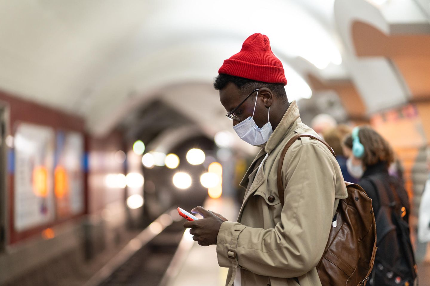 Mann mit Maske steht am Bahnsteig und schaut auf sein Smartphone