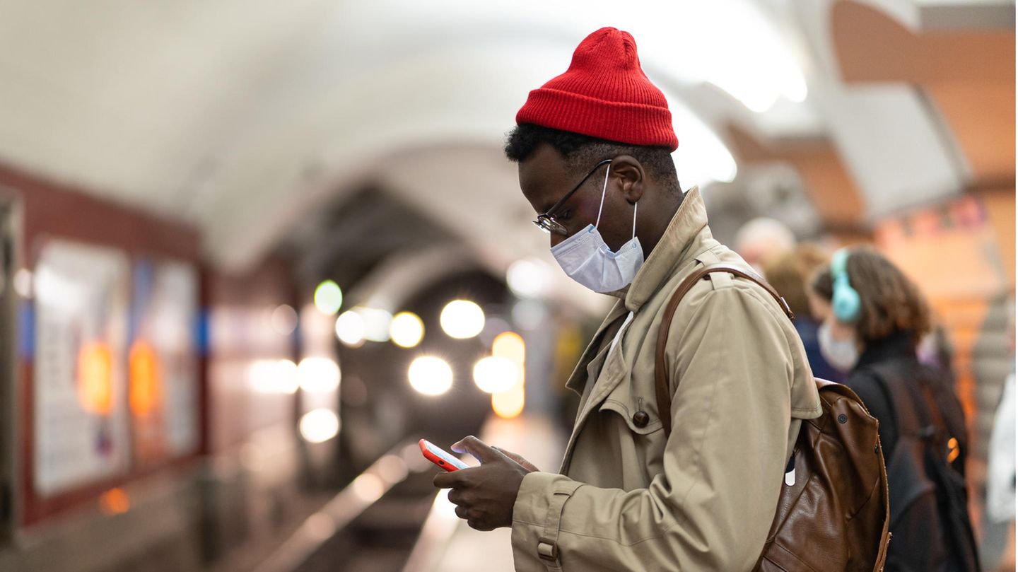 Mann mit Maske steht am Bahnsteig und schaut auf sein Smartphone