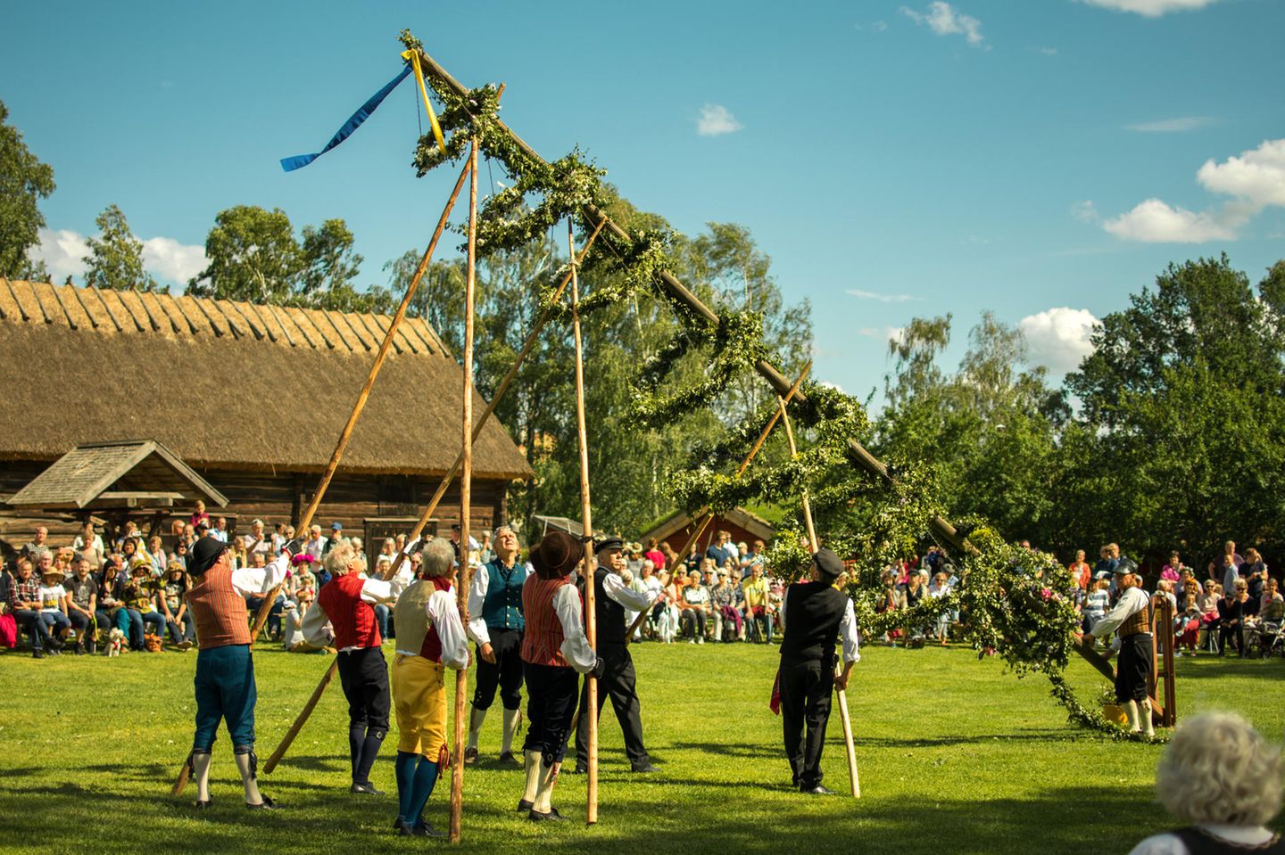 Wenn die Sonne nicht untergeht: Midsommar  Einige Personen in traditioneller Kleidung stellen einen Baum auf einem Platz auf. Um sie herum stehen Zuschauer