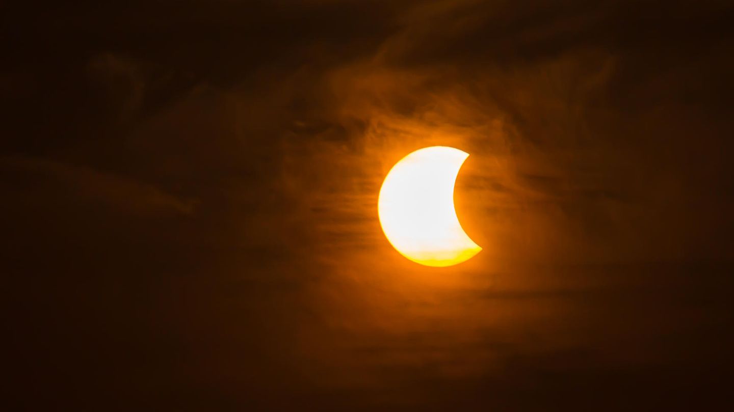 Die Sonne wird von rechts teilweise durch den Mond verdeckt. Rings herum sind Wolken zu sehen.
