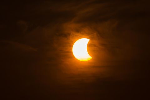 Die Sonne wird von rechts teilweise durch den Mond verdeckt. Rings herum sind Wolken zu sehen.