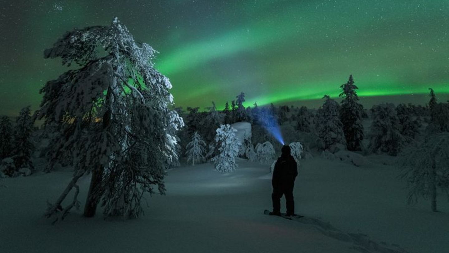 Polarlichter am Himmel und ein Mensch von hinten mit Stirnlampe in verschneiter Landschaft