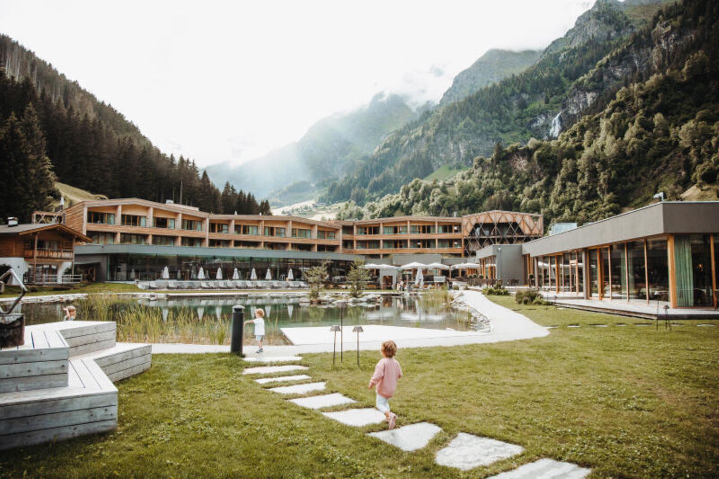 Kategorie Familie: Feuerstein in Südtirol, Italien Kleine Kinder laufen vor dem Hotel entlang, das zwischen den Bergen liegt