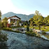 Ein kleiner Fluss fließt im Vordergrund an dem Hotel vorbei. Im Hintergrund sind Berge zu sehen