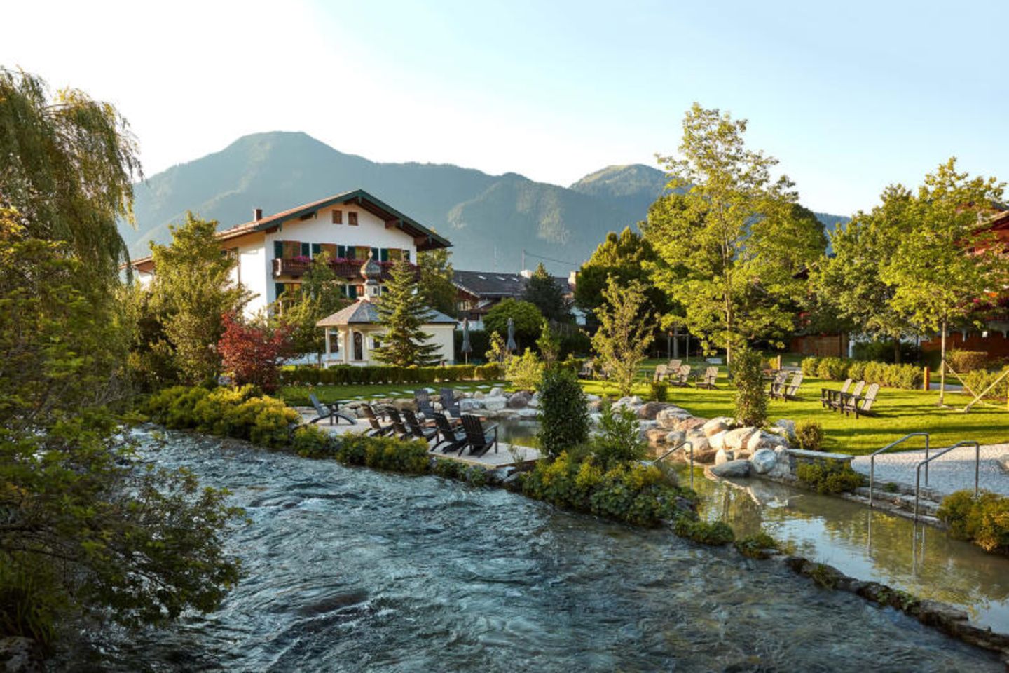 Ein kleiner Fluss fließt im Vordergrund an dem Hotel vorbei. Im Hintergrund sind Berge zu sehen