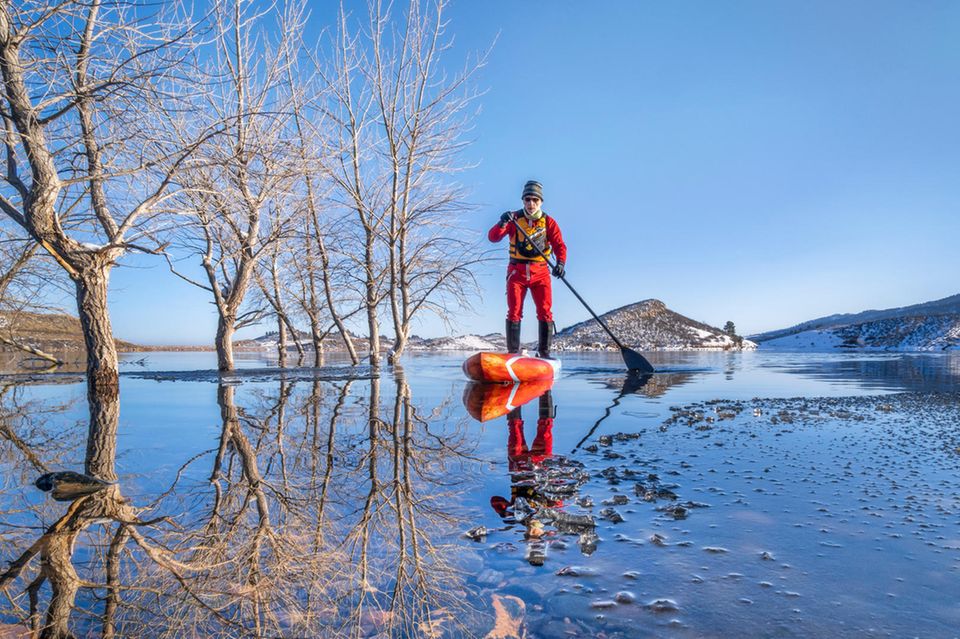 Ein Mann steht in winterlicher Umgebung auf einem SUP-Board auf dem Wasser.