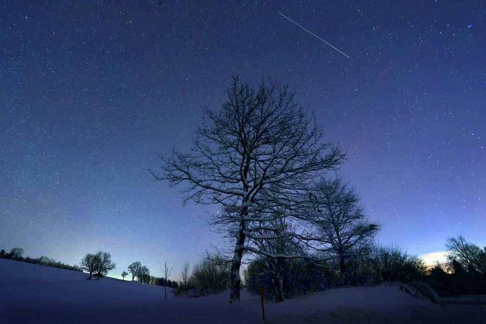 Ein Sternschnuppe verglüht am sternenklaren Himmel über der schneebedeckten Allgäuer Landschaft kurz vor Sonnenaufgang