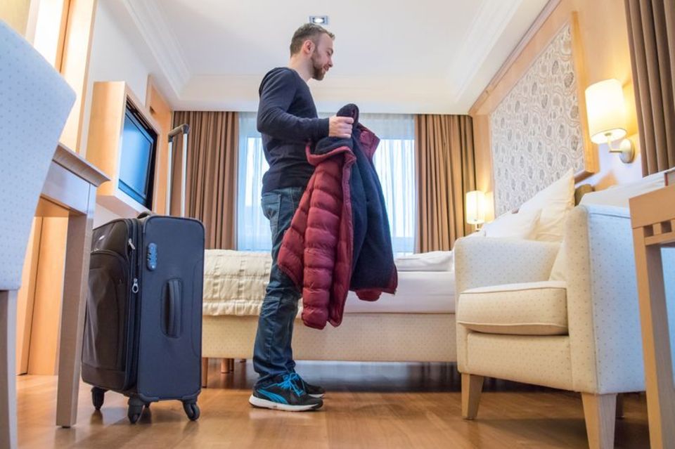 Mann steht mit seinem Koffer in Hotelzimmer und zieht seine Jacke aus