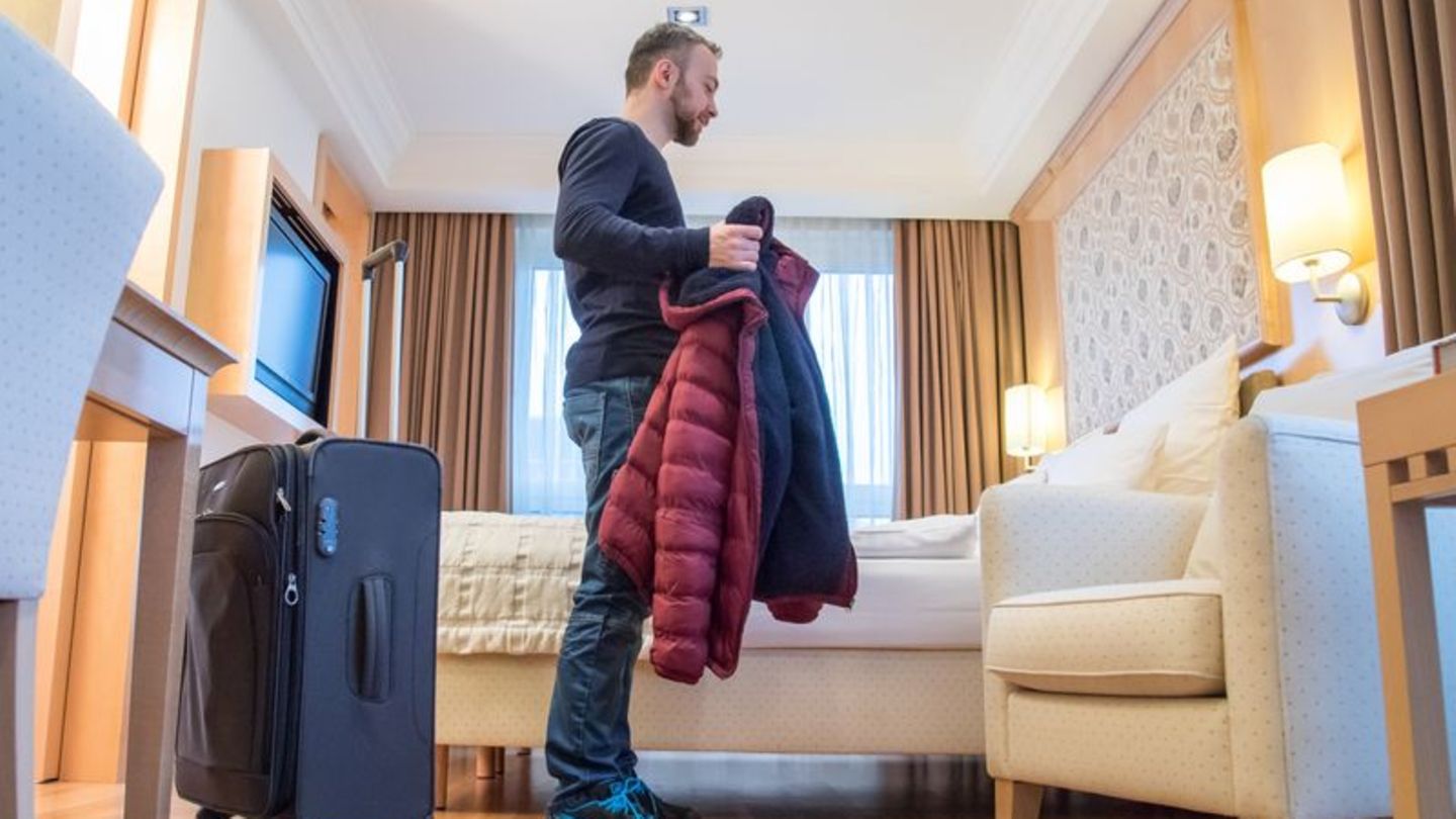 Mann steht mit seinem Koffer in Hotelzimmer und zieht seine Jacke aus