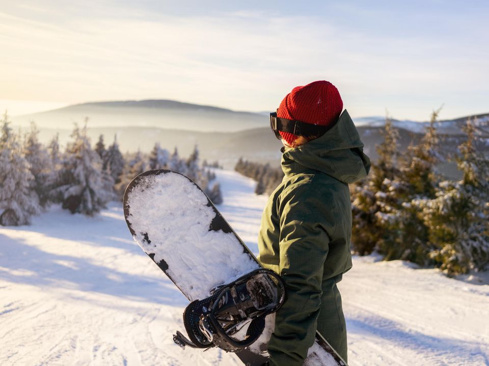Eine Frau mit Snowboard unter dem Arm blickt auf eine Skipiste.