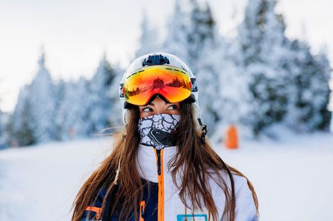 Eine Frau mit Skihelm, Skimaske und anderen Ski Gadgets steht auf einer verschneiten Skipiste.
