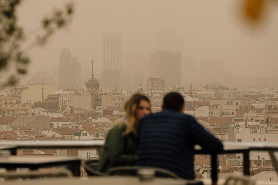 Zwei Menschen sitzen in Madrid auf einer Terrasse, während eine Wolke mit Sahara-Staub über die spanische Hauptstadt zieht. Die