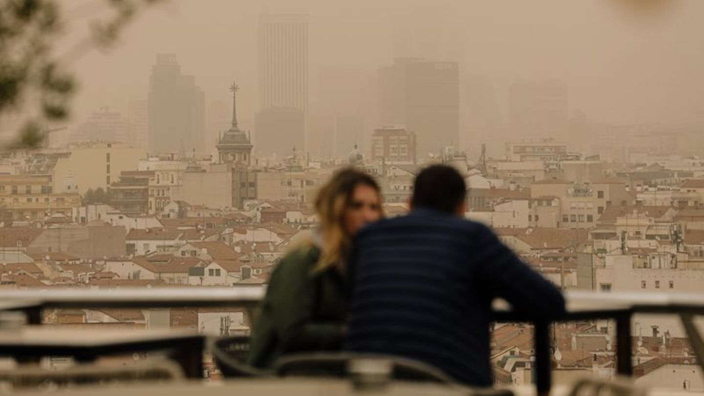 Zwei Menschen sitzen in Madrid auf einer Terrasse, während eine Wolke mit Sahara-Staub über die spanische Hauptstadt zieht. Die