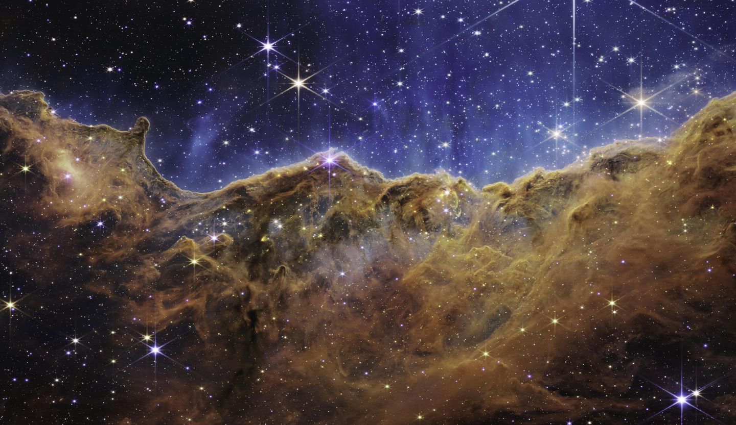 Wie eine Bergkette im letzten Abendlicht leuchtet der Carina-Nebel. Das fast dreidimensional wirkende Bild zeigt eine Region, die in der Astronomie »kosmische Kliffs« heißen. Im blauen Hintergrund schweben Tausende junge, sehr masse-reiche Sterne. Die starke UV-Strahlung und die Sonnenwinde, die von ihnen ausgehen, formen im Rand der braunen Staub- und Gaswolke bizarre Strukturen, die sich teils über mehrere Lichtjahre erstrecken