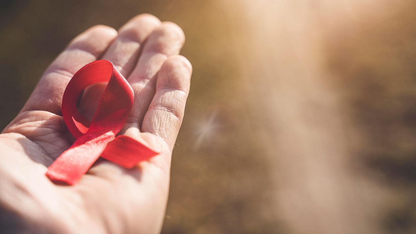 Hand hält eine rote Schleife gegen die Sonne. Symbolbild zum Thema Aids